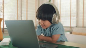 Little Cute Asian Boy in headphones having a video call talking using laptop sitting in the living room at home. Young child boy looking at laptop screen speaking online communication by video webcam