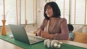 Young Asian Mother stays at home using laptop computer having a video call online conference talking and discussing with the team, working from home while two her son playing on the sofa behind.