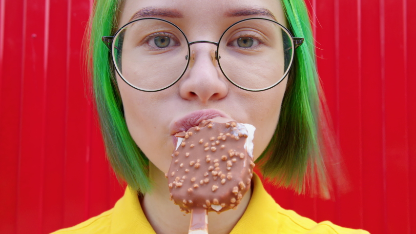 A woman eats an ice cream. Young woman with green hair in yellow t-shirt bites an eskimo pie. Close up. Red background | Shutterstock HD Video #1064567410