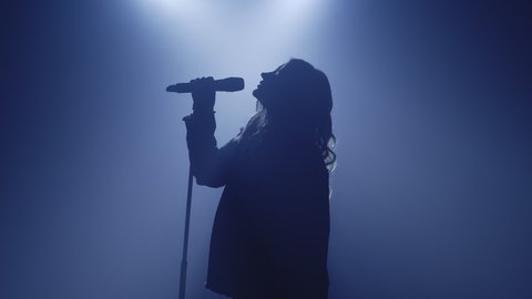 Backlit silhouette of singer vocalist girl standing in dark nightclub disco studio and start singing performance. Light appears and illuminates the woman. Isolated on black background. Slow motion
