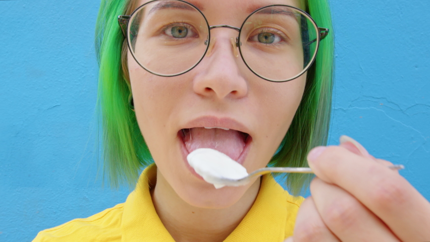 A cheerful woman in glasses eating a spoon of yogurt. A young woman with green hair against blue background. Concept of healthy lifestyle | Shutterstock HD Video #1064569801