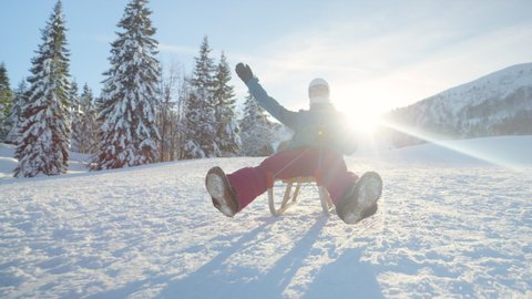 SLOW MOTION, CLOSE UP, LOW ANGLE, LENS FLARE: Young woman on an active winter vacation in Slovenian mountains goes sledding on a sunny day. Female tourist is enjoying her winter holiday by sleighing.