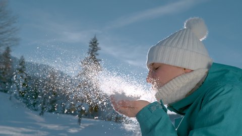 SUPER SLOW MOTION, LENS FLARE, CLOSE UP, DOF: Playful female tourist blowing snowflakes into the bright winter sunshine. Smiling young Caucasian woman blows away handful of freshly fallen powder snow.
