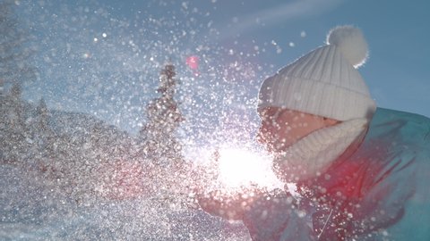 SLOW MOTION TIME WARP, LENS FLARE, CLOSE UP, DOF: Smiling young Caucasian woman blows away a handful of fresh powder snow. Playful female tourist blowing snowflakes into the bright winter sunshine.