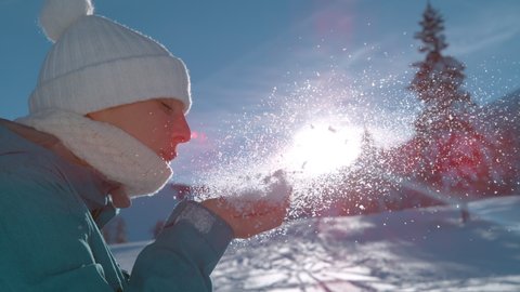 SUPER SLOW MOTION, CLOSE UP LENS FLARE, DOF: Cheerful young woman holds up a handful of snow and blows it away. Snowflakes sparkle in the winter sunshine as female tourist blows into hand full of snow
