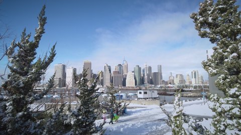 New York, USA - Dec 17, 2020: NYC Manhattan skyline from Brooklyn Bridge Park on a Snow day during the holidays and coronavirus pandemic (covid-19). Snow and trees. Family and jogger.