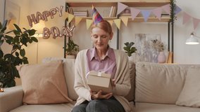 Medium pov of senior woman sitting on couch in festively decorated living room opening box with birthday present feeling happy and surprised having online birthday party