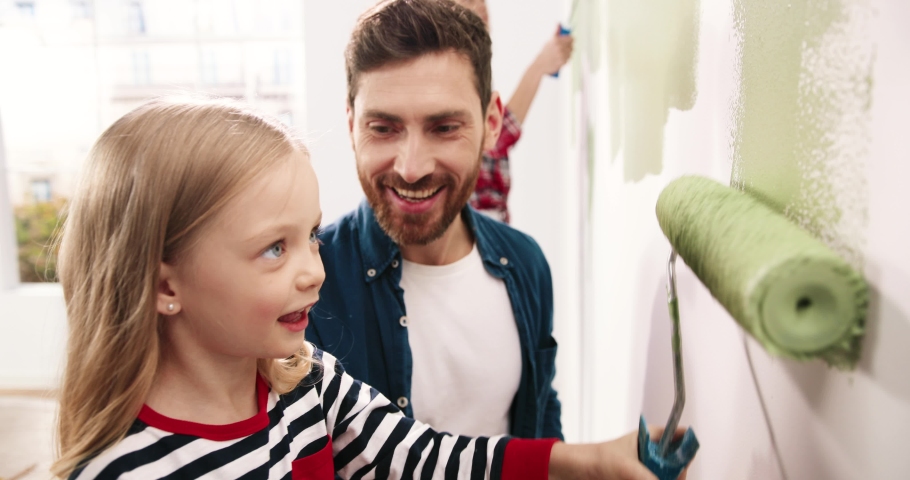 Joyful young Caucasian family painting room. Dad teaching little adorable daughter to paint wall using roller brush. Happy child girl helping parents to renovate house. Home repair concept. Close up Royalty-Free Stock Footage #1064589436
