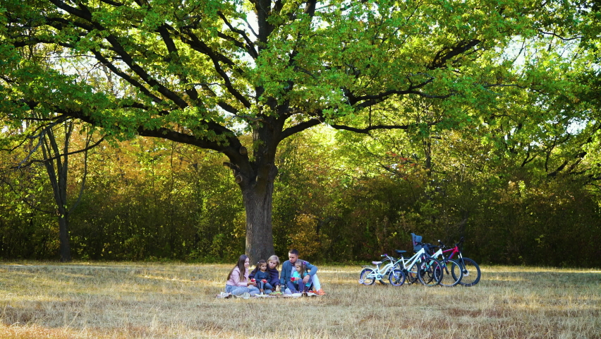 Family sitting under big oak tree and having picnic, bicycles standing not far from them. Parents and children relaxing after biking in park. Concept of bonding