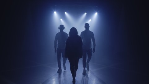 Backlit silhouettes of singer vocalist girl, saxophonist sax, dj man walking forward in dark musician nightclub disco for starting concert on stage. Light appears and illuminates musical group band