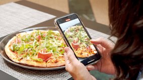 Close-up of female hands holding smartphone and making photo or video of delicious pizza in a restaurant. Blogging, technology and social media concept.