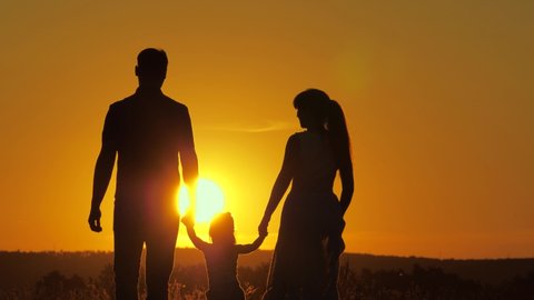 Happy family, little daughter is jumping, holding hands of dad and mom in park in sun. The child plays with dad and mom on field in light of sunset. Walk with a small child in nature. childhood