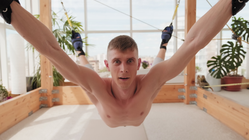 Medium close-up portrait of slim sportsman lifted in air parallel to floor attached by rope to special fasteners smiling at camera trying unusual stretching simulator
