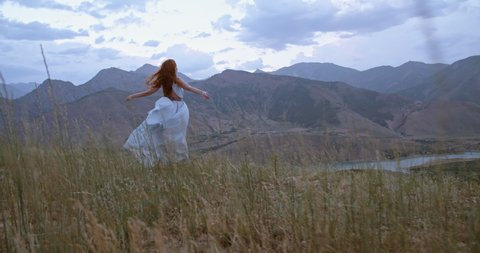 Young beautiful girl with red hair wearing white dress walking on top of a mountain facing wind blowing her hair and dress - freedom, adventure, harmony 4k footage