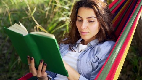Close up Portrait of a Young Adult Girl Lying in a Hammock and Reading a Book on Nature in the Park. Pretty Woman Rest and Relax with a Book in hand. A Student Hipster Learns Outdoors.