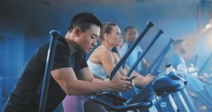 Asian cheerful man trainingin sports club on eliptical bike using smartphone recording video messages for girlfriend rejoicing. Sportsman. Busy lifestyle. Nomophobia.