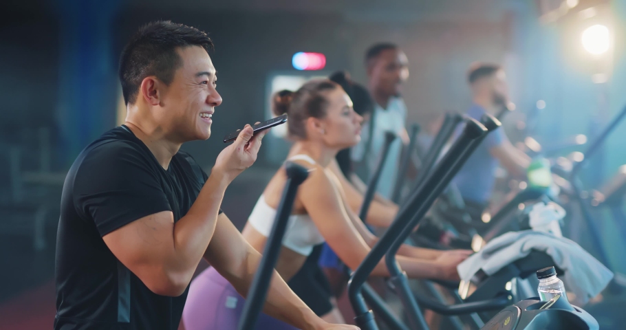 Asian handsome young positive man chatting messages using smartphone recording voice audio message while workingout with team sports in modern gym.