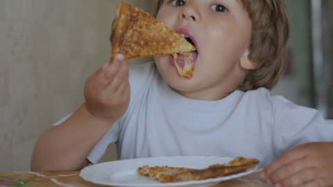 Child Eat Pizza. Little Hungry Boy Eating Tasty Italian Pizza From Plate Sitting Home at Table in Kitchen. Unhealthy Nutrition Fast Food. Portrait of a Kid Eating Lunch. Face Young Little Boy Closeup.