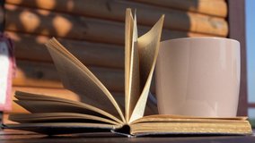 Closeup view 4k video of old open paper book laying on wooden table on sunny terrace and mug with steaming hot drink