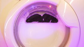 White washing machine washes dirty colorful clothes. Washing clothing in domestic washing machine. Close up video of spinning drum washing machine.