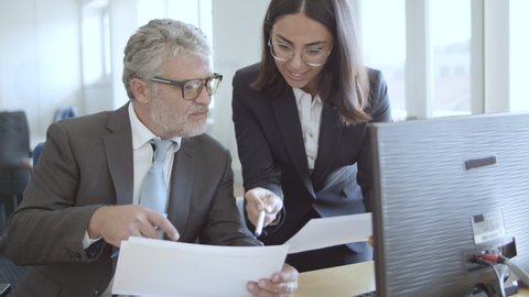 Friendly positive female assistant in suit and glasses explaining document details to male boss, pointing pen at papers. Crane shot. Business and cooperation concept