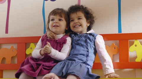 Caucasian and hispanic female preschoolers sitting on bench in kindergarten, playing and smiling. Portrait of two little friends laughing at school