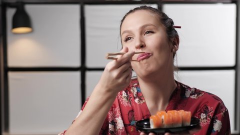 Woman eating sushi. Happy smiling woman dressed in red dress in Asian style eats sushi rolls from black porcelain plate, enjoys meal. Copy space composition