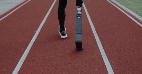 Backside view of unknown disabled male person with prosthetic running blades walking at sports field. Amputee male runner getting ready. Concept of motivational sports footage