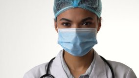 Health and care. Attractive young female doctor wearing medical mask blinking while looking at camera, posing isolated over white background. Healthcare and medicine concept