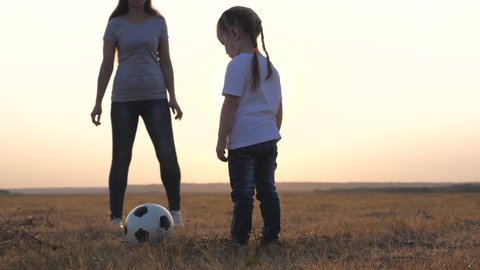 A little daughter kicks a soccer ball in field, ball rolls towards her mother. Family at sunset in meadow cheerfully plays football. Happy family of children and mom having fun playing ball in park.