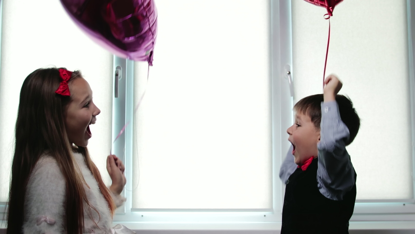 Happy dressed-up children, a boy and a girl, with helium balloons in the shape of a heart in their hands, laugh and hug. Concept of Valentine's Day, 14th February Royalty-Free Stock Footage #1064649871