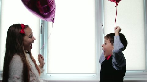 Happy dressed-up children, a boy and a girl, with helium balloons in the shape of a heart in their hands, laugh and hug. Concept of Valentine's Day, 14th February