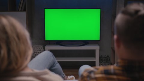 Family Couple Watches Green Screen TV Mockup Sitting on Couch in Living Room Together. Rear View on Casual People who Watching TV Green Screen in Domestic Cinema. Looking TV Show or News in Home Rest