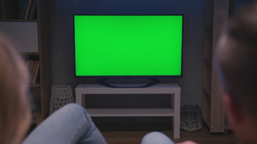 Family Couple Watches Green Screen TV Mock-up Sitting on Couch in Living Room Together. Rear View on Casual People who Watching TV Green Screen in Domestic Cinema. Looking TV Show or News in Home Rest