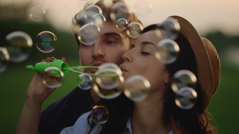 Portrait of playful couple blowing soap bubbles on meadow. Closeup happy man and woman having fun at romantic date outdoors. Young couple throwing bubbles at weekend in summer park