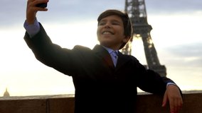 A smiling teenage boy in a black suit is taking a selfie on the phone on the background of the Eiffel tower at the dawn, Paris, France