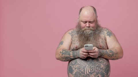 Shocked fat pudge obese chubby overweight bearded man has tattooed naked bare big belly isolated on pink background studio. People lifestyle concept. Using mobile cell phone point index finger aside