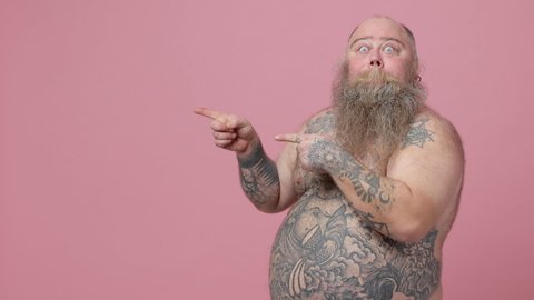 Excited fat pudge obese chubby overweight bearded man has tattooed naked big belly isolated on pink background. People lifestyle concept. Pointing camera aside doing winner gesture showing thumbs up