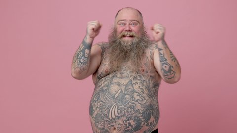 Side view of excited fat pudge obese chubby overweight bearded man has tattooed naked big belly isolated on pink background. People lifestyle concept. Turn around camera doing winner gesture dancing