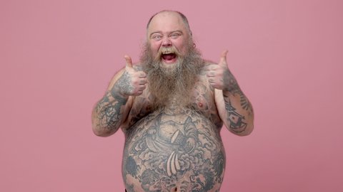Excited crazy fat pudge obese chubby overweight bearded man has tattooed naked bare big belly isolated on pink background studio. People lifestyle concept. Showing thumbs ip like gesture screaming