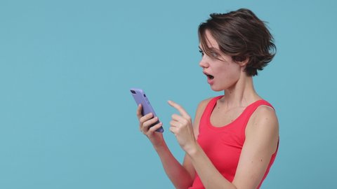 Side view of shocked young woman 20s in pink tank top posing isolated on blue background in studio. People lifestyle concept. Point index finger on mobile cell phone browsing say wow showing thumb up