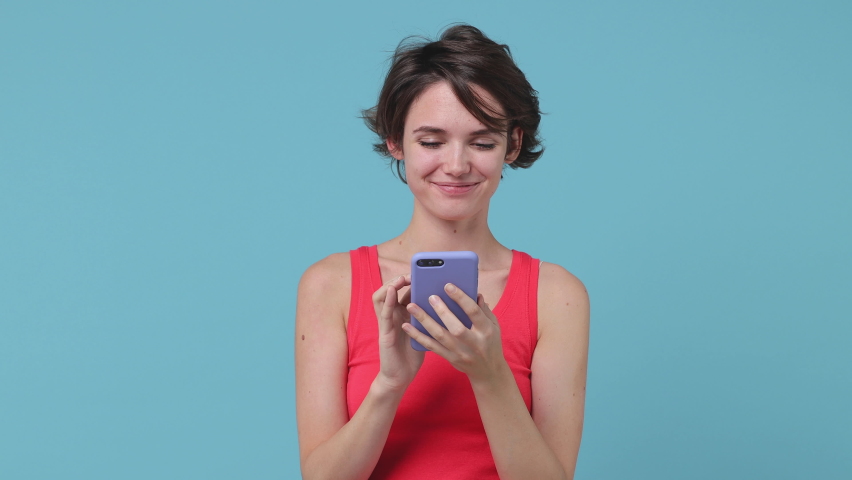 Excited joyful brunette young woman 20s years old in pink tank top posing isolated on pastel blue background studio. People lifestyle concept. Talking on mobile cell phone doing winner gesture say yes