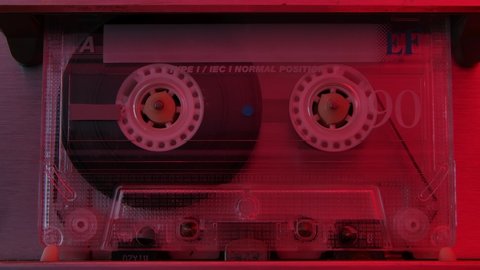 Retro compact audio cassette playing extreme close up. Vintage music cassette playing back in the player illuminated by red neon lights. Slow motion.