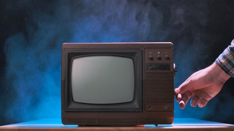 Man tries to turn on the old TV, knocks on it, scattering dust particles. Vintage television against a smoky studio background with blue neon lights. Male hands close up. Slow motion.
