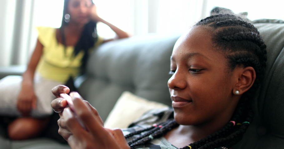 Black girl playing game on phone at home couch, sister watching sibling use smartphone device.