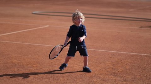 A small child learns to hit a ball with a racket. Learning to play tennis on a sports field with a clean outdoor. Summer activity.