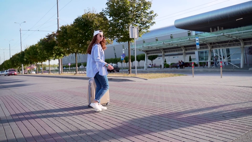 Cheerful Young Woman in Summer Cap and Sunglasses Walking Near Airport with Suitcase. Happy Lady with dark hair Enjoying Adventure Time. Traveling Hipster Girl Student Tourist 