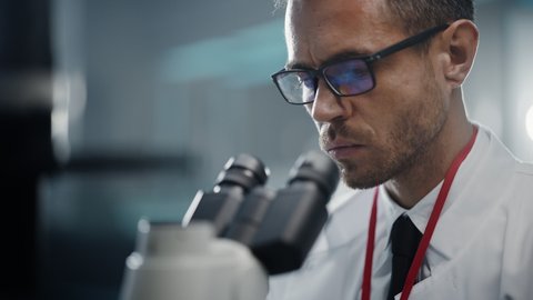 Medical Development Laboratory: Portrait of Caucasian Scientist Looking Under Microscope, Analyzes Petri Dish Sample. Big Pharmaceutical Lab doing Medicine, Biotechnology, Microbiology, Drugs Research
