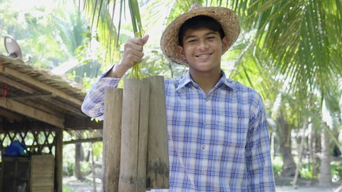 A smiling young Asian male farmer shows a bamboo cane with a handle and a handle to collect sugar from coconuts in the garden to bring to a boil and simmer to precipitate into coconut sugar cubes.