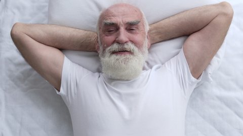 Glad greyhaired pensioner stretching in bed, waking up on comfortable pillow